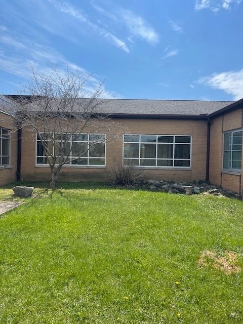 The exterior of a single-story brick school building with large windows, a small bare tree, and a rock garden border along the foundation, surrounded by a green lawn under a blue sky.