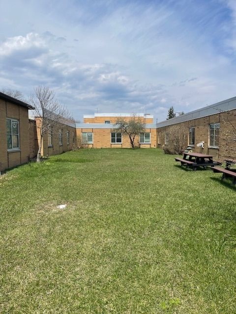 A grassy courtyard enclosed by single-story brick school buildings, with picnic tables on the right side, bare trees, and a partly cloudy sky overhead.