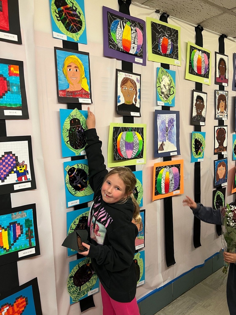 A view of an art exhibition hallway with vibrant student paintings and drawings covering the walls on both sides, with a person visible in the distance.