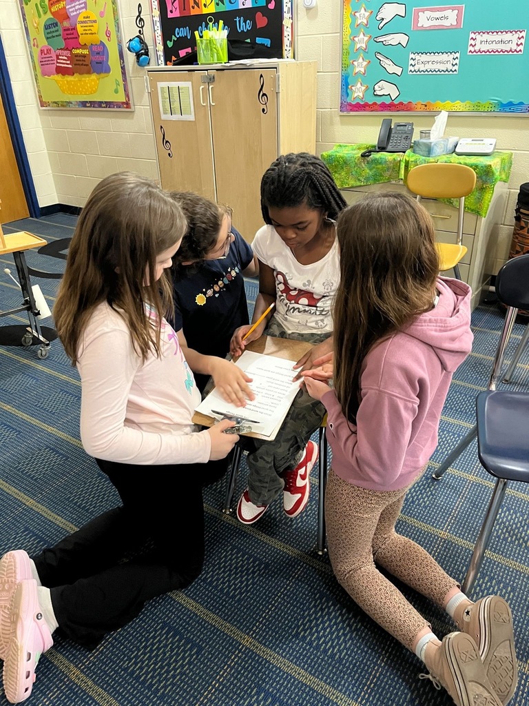 A small group of elementary-aged children sit on the floor together, looking at a paper on a clipboard.