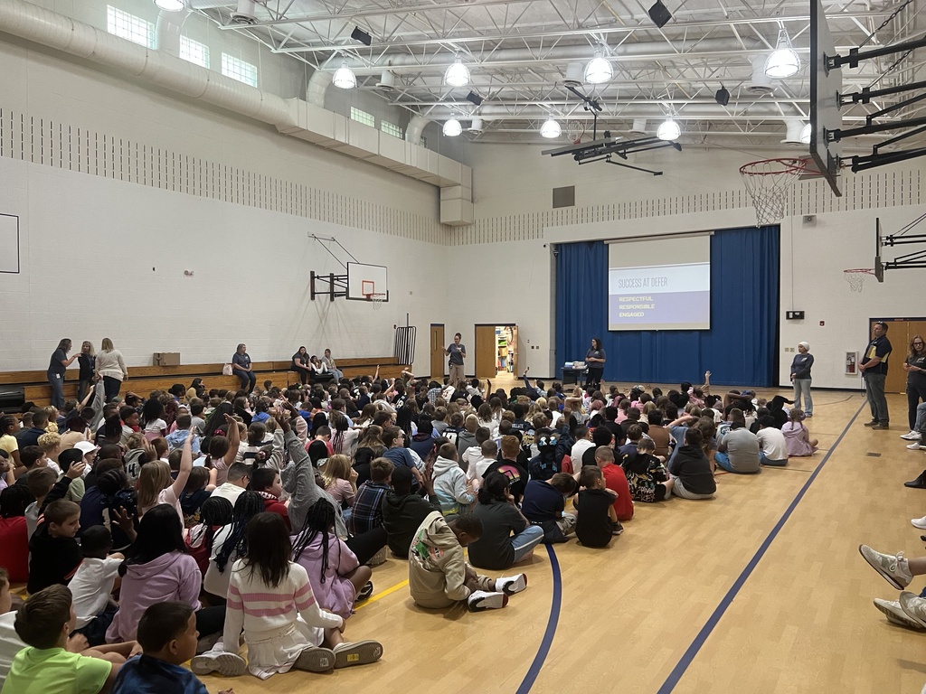All HDI students sitting on the floor of the gym while staff talk to them at a podium with a projection screen behind them. 