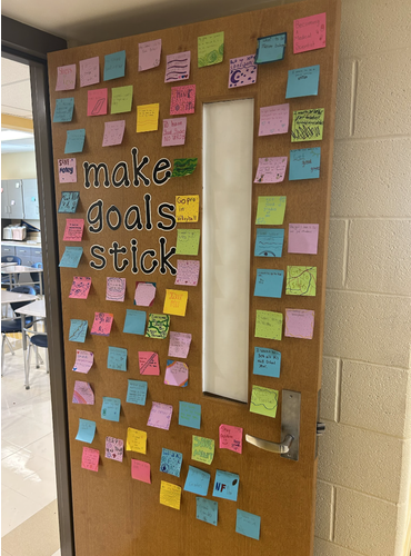 Photo of an open door to Mrs. Speakman's classroom. The door has big letters in black that says "Make Goals stick" Surrounding those words are a bunch of different post-it notes in a variety of colors featuring student goals for the school year. 