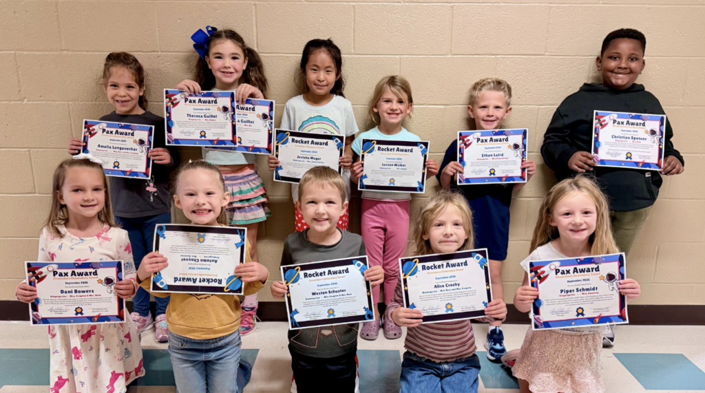 A group of kindergarten students holding certficates. There are six students standing in the back row against a wall and then kneeling in the front row are five students. 