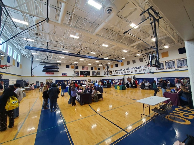 A wide-angle view of a career fair filling the gymnasium of a school whose wall reads "Home of the Rockets." Students browse dozens of employer booths lining the court, with championship banners, an American flag, and basketball hoops visible overhead.