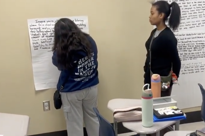 Students are standing in front of a wall in a classroom. One is writing on an oversized piece of paper and one is watching.