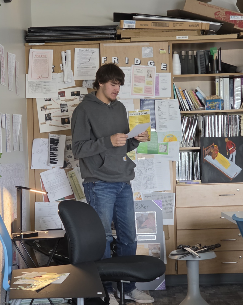 A senior in high school wearing jeans and a gray hoodie standing up and giving a presentation.