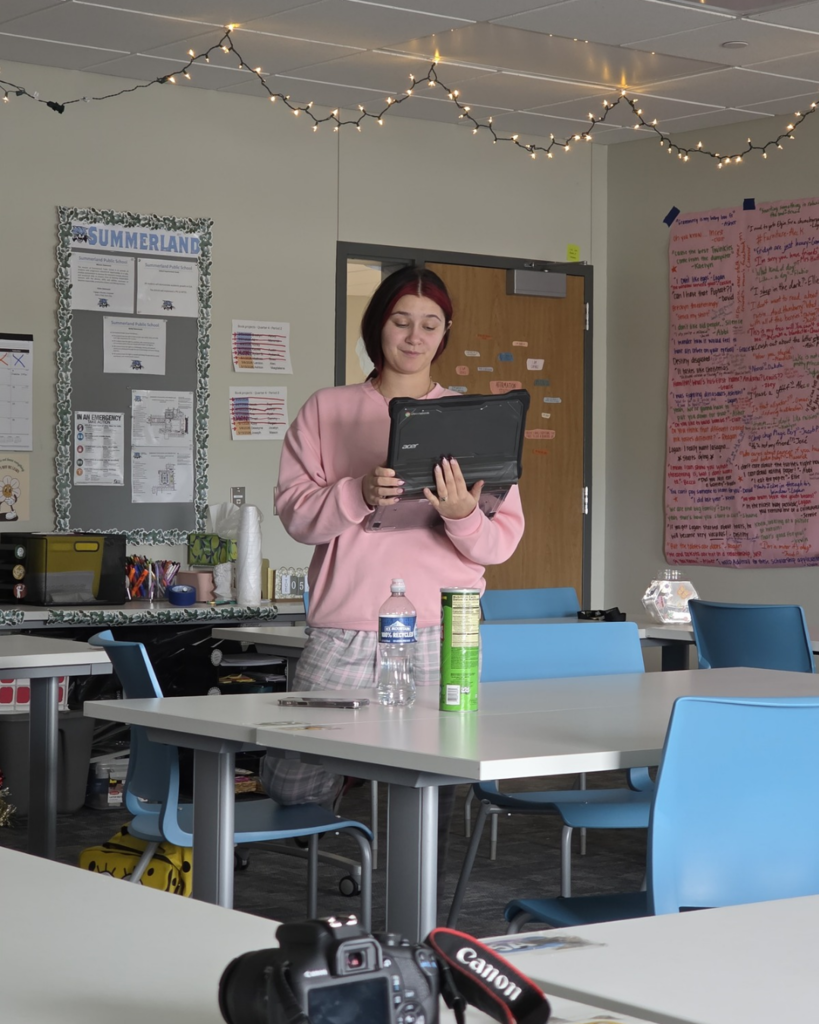 A senior girl in high school with a pink sweatshirt standing up and giving a presentation from a computer. 