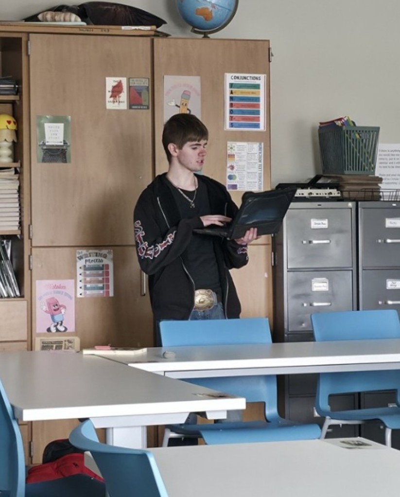 A senior boy in high school wearing jeans and a black shirt, standing up and giving a presentation from a computer.