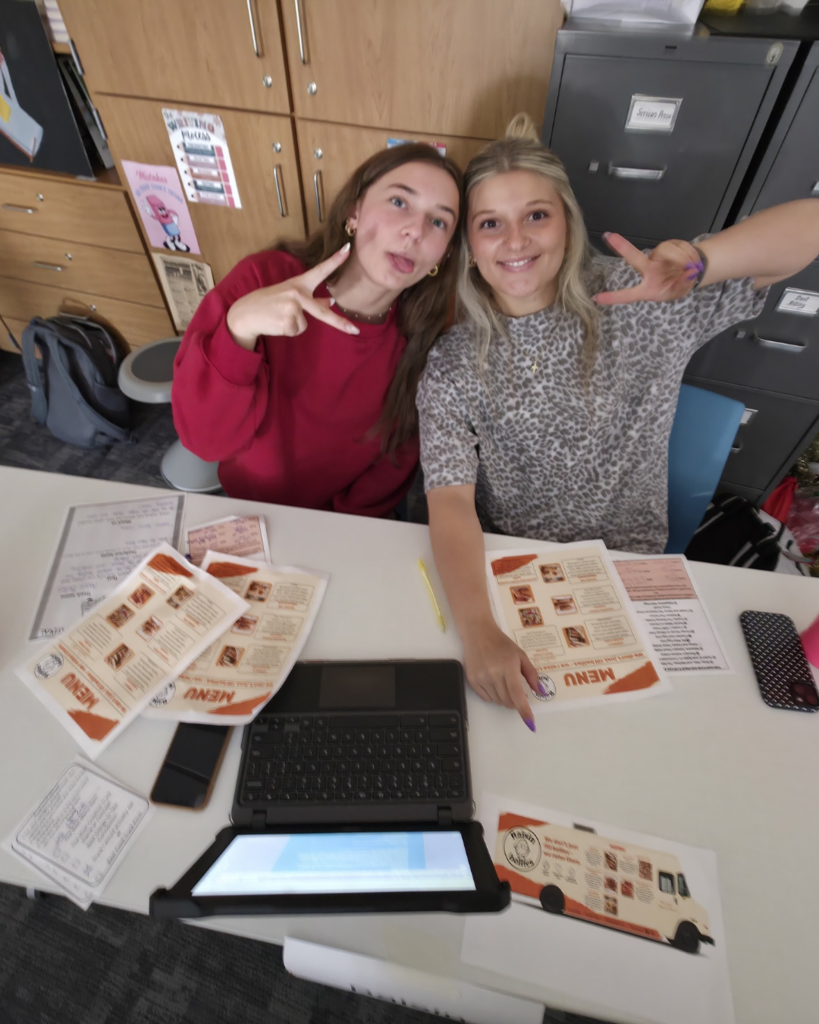 Two girls pose for a picture after they presented their food truck called "Raisin Bellies." Images of their menu and their truck can also be seen on the table in front of them.