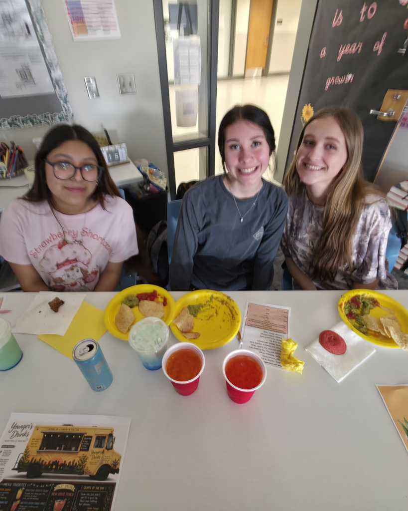 Three girls pose for a picture and smile in front of their truck "Younger Drinks" with plates full of chips and drinks at their table.