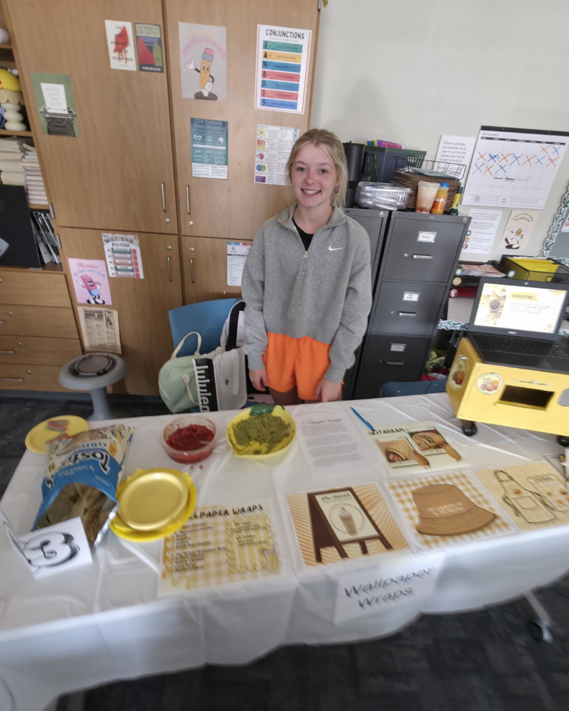A girl wearing gray stands behind her food truck table called "Wallpaper Wraps" where she is showing off her yellow merchandise and has chips and dip for people to try.