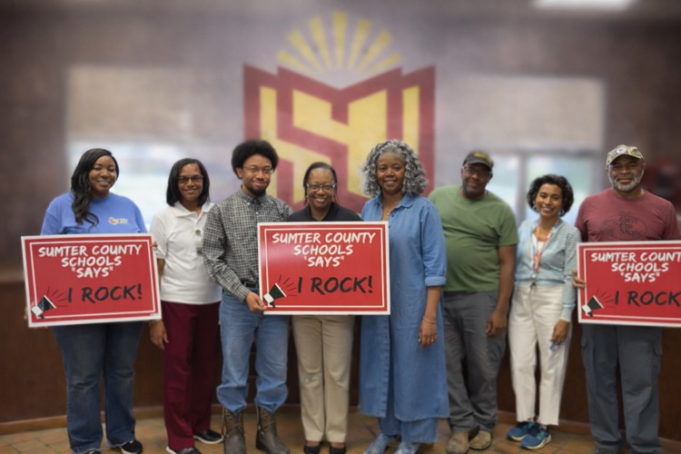 A group of Sumter County Schools employees stand together smiling while holding signs that read “Sumter County Schools Says I Rock!” 