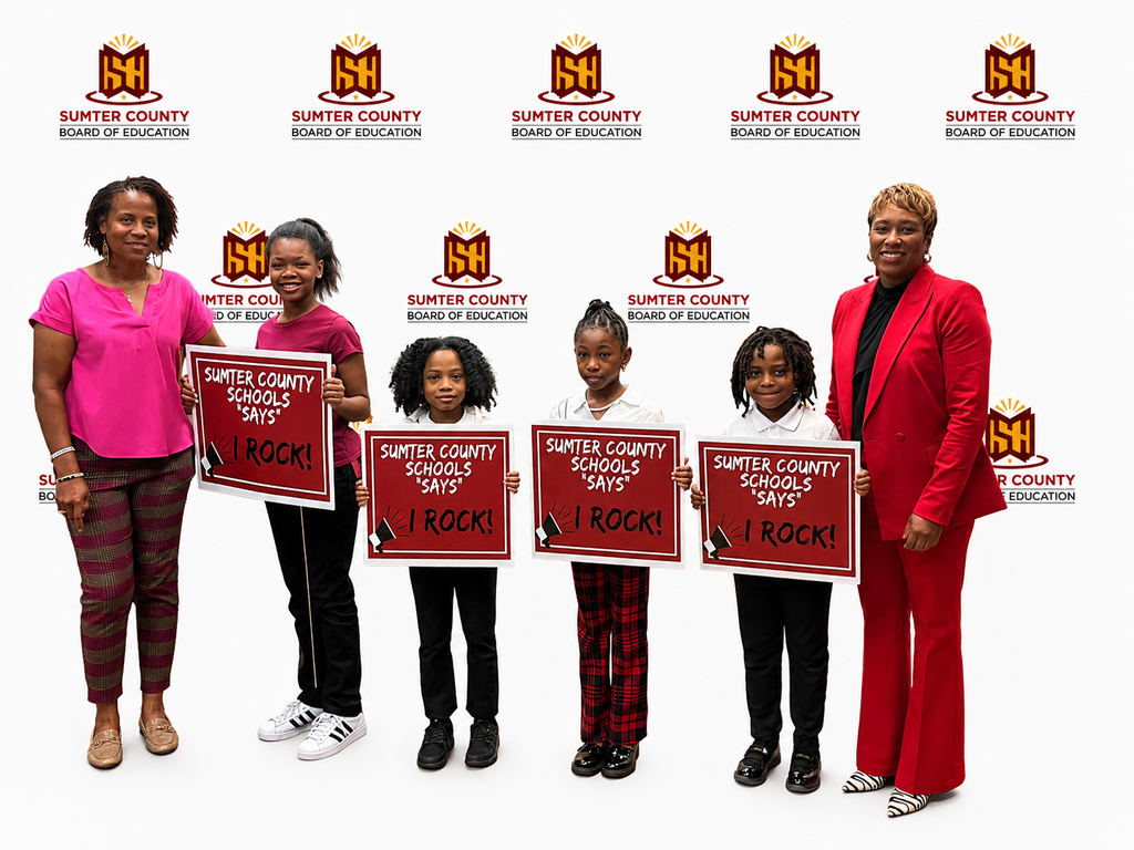 A group of Sumter County Schools students, faculty, and staff members stand together during a Board of Education meeting while holding red “Sumter County Schools Says I ROCK!” signs. Honorees included Child Nutrition Program workers, custodians, bus drivers, secretaries, paraprofessionals, auxiliary teachers, classroom teachers, support personnel, and students recognized for their dedication and service. Superintendent Dr. Marcy Burroughs is pictured with several honorees during the recognition ceremony. The meeting takes place inside the Sumter County Board of Education boardroom.