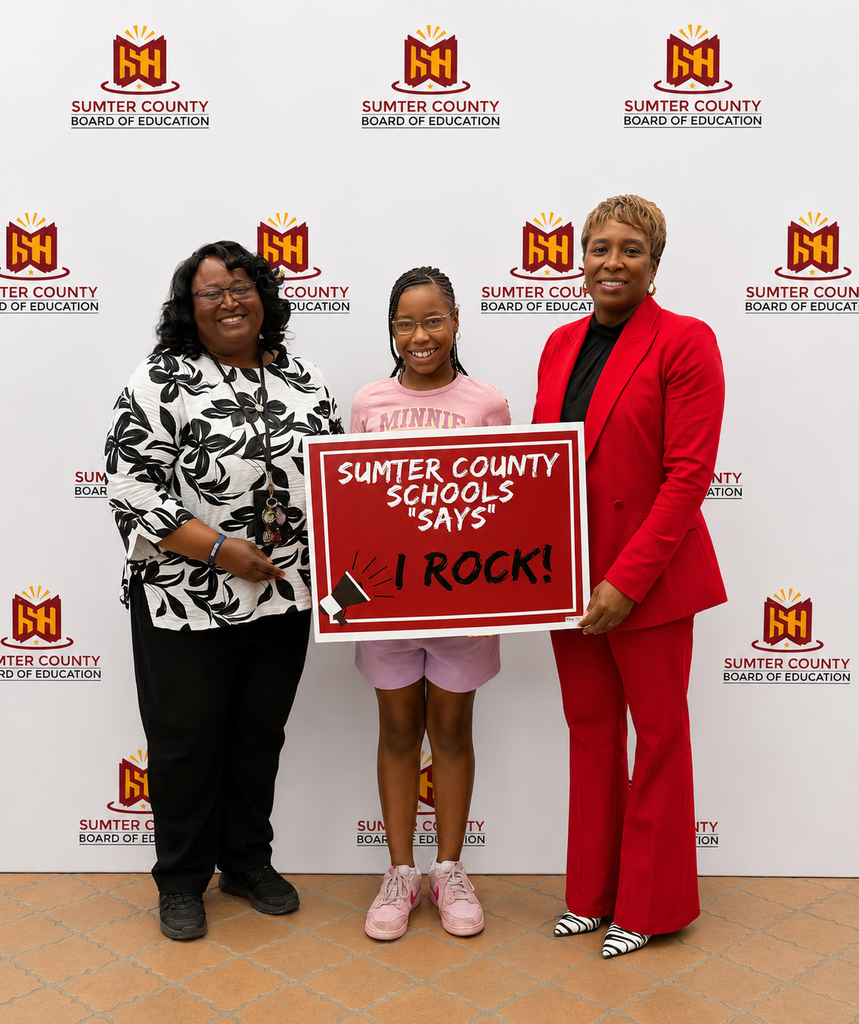 A group of Sumter County Schools students, faculty, and staff members stand together during a Board of Education meeting while holding red “Sumter County Schools Says I ROCK!” signs. Honorees included Child Nutrition Program workers, custodians, bus drivers, secretaries, paraprofessionals, auxiliary teachers, classroom teachers, support personnel, and students recognized for their dedication and service. Superintendent Dr. Marcy Burroughs is pictured with several honorees during the recognition ceremony. The meeting takes place inside the Sumter County Board of Education boardroom.