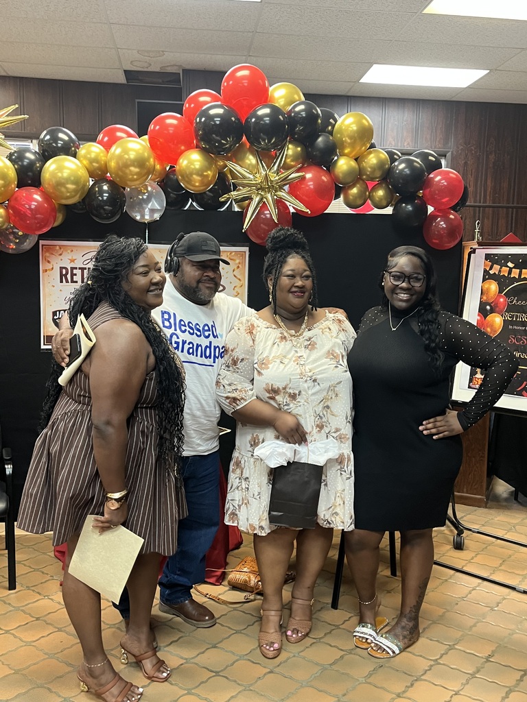 A retirement celebration photo featuring two women standing together in front of a black backdrop decorated with red, gold, and black balloons and gold star accents. One woman is wearing a cream floral dress with tan heels, and the other is wearing a red cardigan with black pants and white shoes. Behind them is a retirement-themed sign for Sumter County Public Schools, creating a festive and celebratory atmosphere.