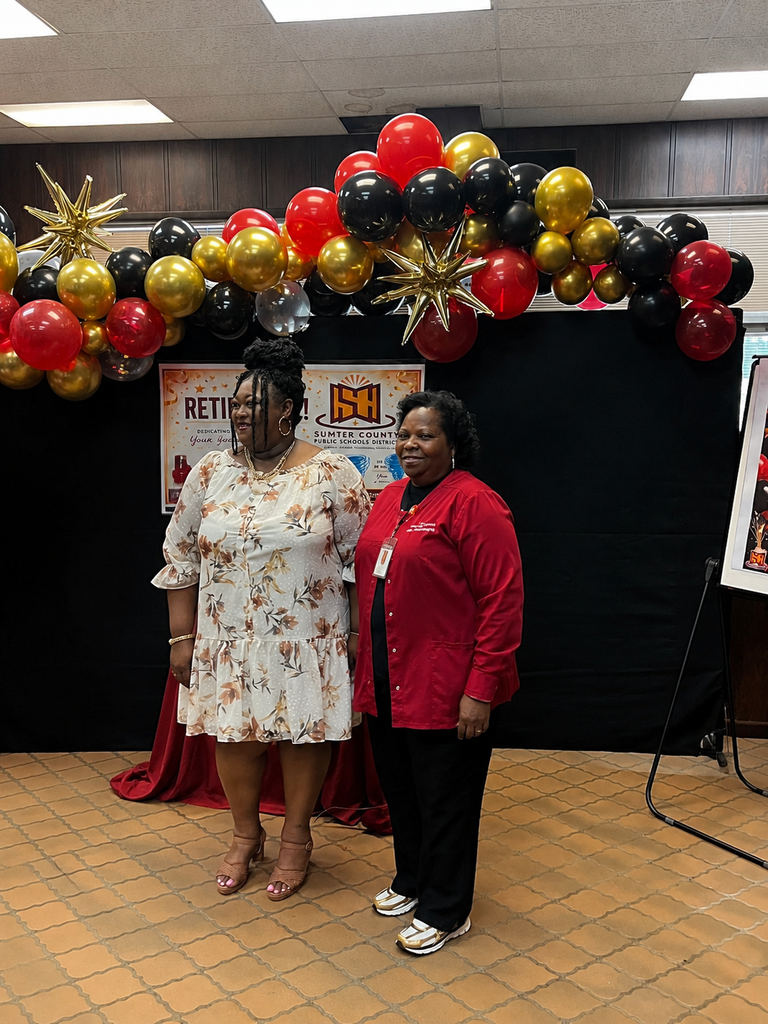 A retirement celebration photo featuring two women standing together in front of a black backdrop decorated with red, gold, and black balloons and gold star accents. One woman is wearing a cream floral dress with tan heels, and the other is wearing a red cardigan with black pants and white shoes. Behind them is a retirement-themed sign for Sumter County Public Schools, creating a festive and celebratory atmosphere.