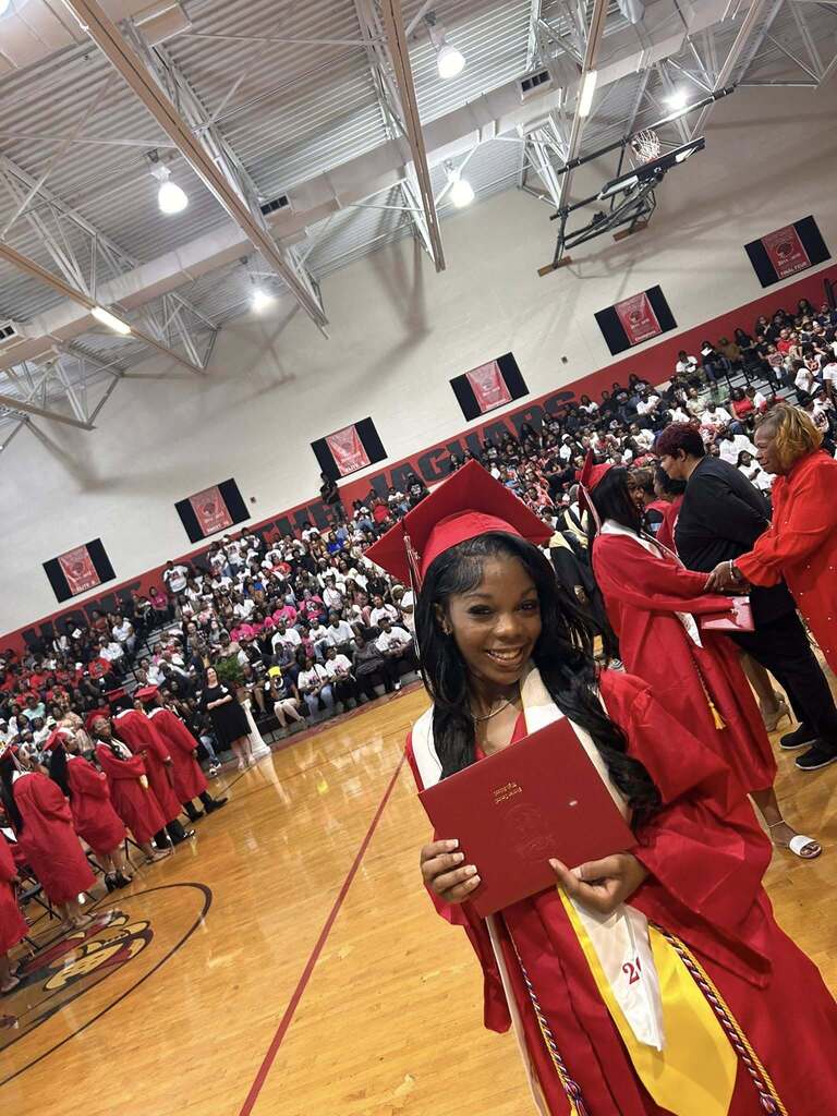 A group of Sumter Central High School seniors dressed in red graduation caps and gowns with white stoles stand together in a school hallway before their graduation ceremony. The graduates are gathered around a staff member in academic regalia, smiling and preparing to process into the ceremony.