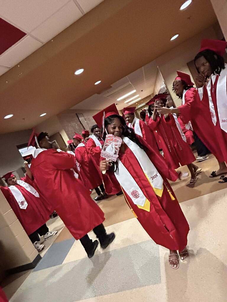 A group of Sumter Central High School seniors dressed in red graduation caps and gowns with white stoles stand together in a school hallway before their graduation ceremony. The graduates are gathered around a staff member in academic regalia, smiling and preparing to process into the ceremony.