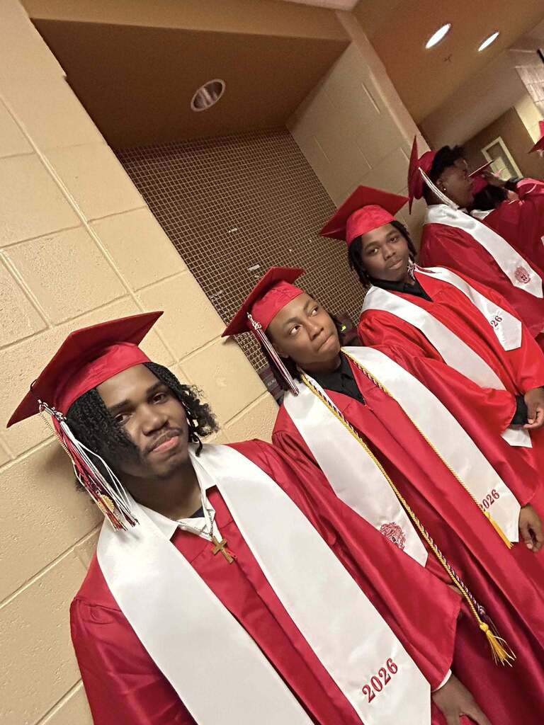 A group of Sumter Central High School seniors dressed in red graduation caps and gowns with white stoles stand together in a school hallway before their graduation ceremony. The graduates are gathered around a staff member in academic regalia, smiling and preparing to process into the ceremony.