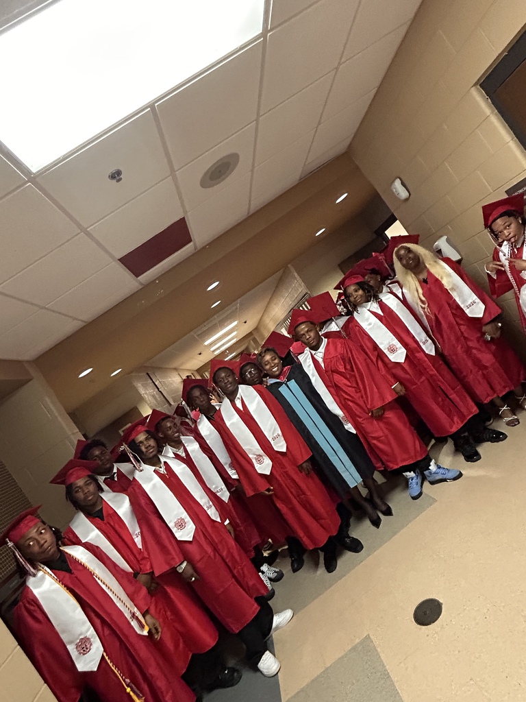 A group of Sumter Central High School seniors dressed in red graduation caps and gowns with white stoles stand together in a school hallway before their graduation ceremony. The graduates are gathered around a staff member in academic regalia, smiling and preparing to process into the ceremony.