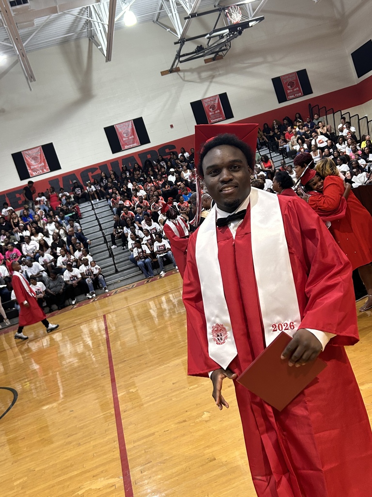 A group of Sumter Central High School seniors dressed in red graduation caps and gowns with white stoles stand together in a school hallway before their graduation ceremony. The graduates are gathered around a staff member in academic regalia, smiling and preparing to process into the ceremony.