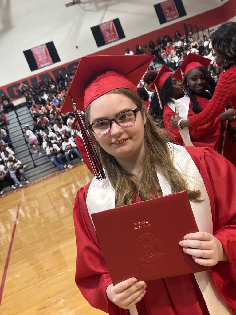 A group of Sumter Central High School seniors dressed in red graduation caps and gowns with white stoles stand together in a school hallway before their graduation ceremony. The graduates are gathered around a staff member in academic regalia, smiling and preparing to process into the ceremony.