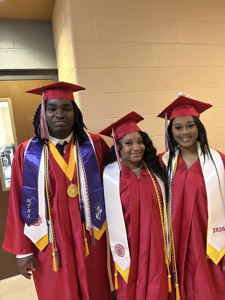 A group of Sumter Central High School seniors dressed in red graduation caps and gowns with white stoles stand together in a school hallway before their graduation ceremony. The graduates are gathered around a staff member in academic regalia, smiling and preparing to process into the ceremony.