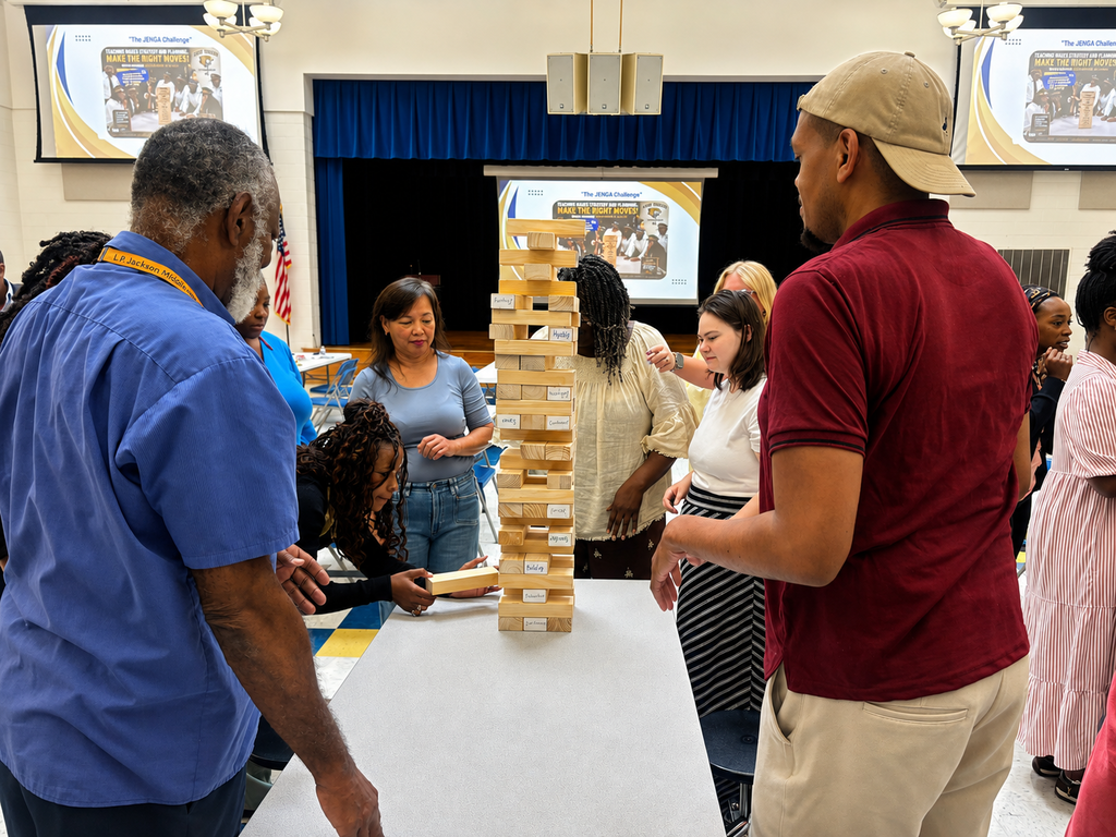Surry County Public Schools staff participate in a team-building Jenga activity during New Employee Orientation, working together around a large wooden block tower in the school auditorium.