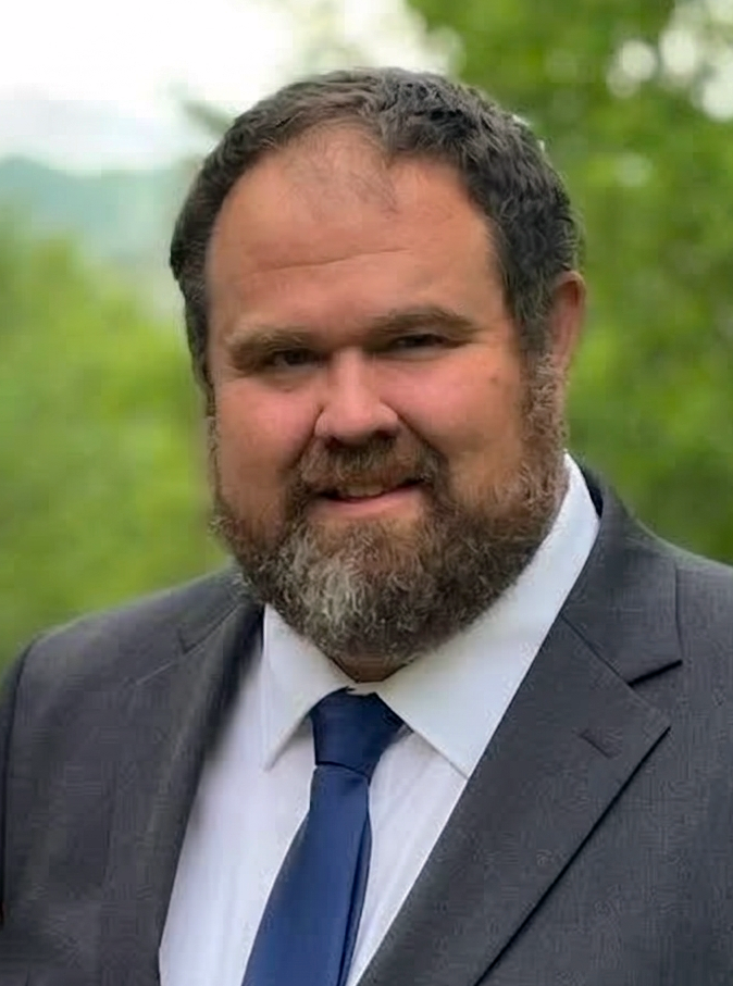 Gemini said The image shows a headshot of Brandon Sutton. He is a middle-aged man with short brown hair and a full, well-groomed beard and mustache. He is smiling warmly and looking directly at the camera. He is dressed professionally in a dark charcoal gray suit jacket, a crisp white collared shirt, and a solid navy blue tie. The background is a soft, out-of-focus lush green, suggesting an outdoor setting with trees or foliage.