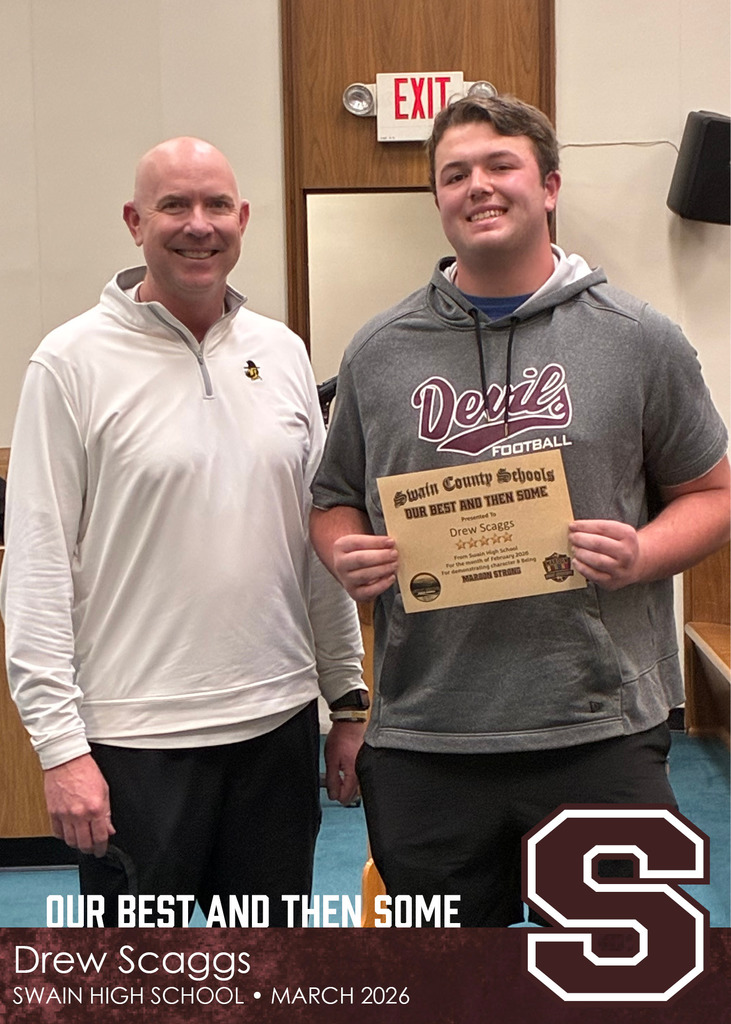 A smiling adult man stands next to a teenage boy wearing a “Devils Football” hoodie. The boy holds a certificate from Swain County Schools recognizing him for character, while both pose indoors in front of a wooden door with an exit sign.