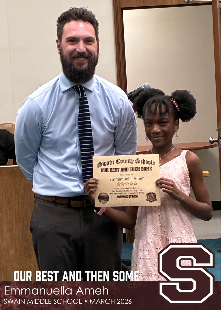 A man in a dress shirt and striped tie stands beside a young girl in a light pink dress. The girl smiles while holding a Swain County Schools certificate, and they pose together in a meeting room.