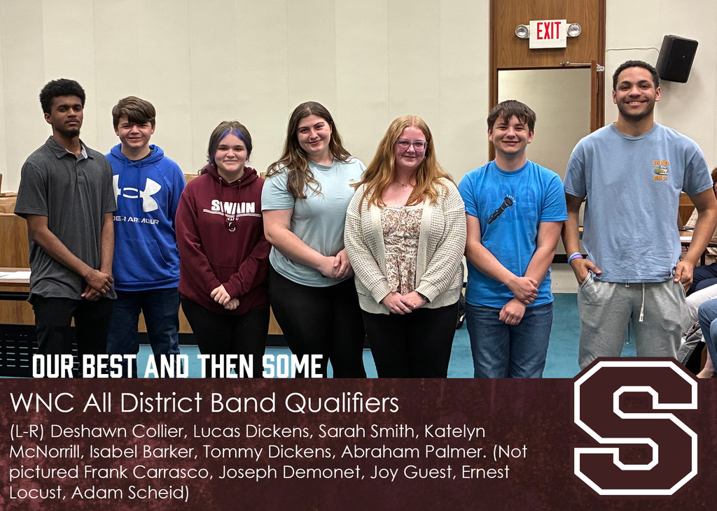 Seven teenagers stand in a row indoors, smiling at the camera in a meeting room with wood paneling and an exit sign behind them.