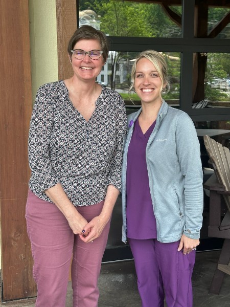 Two women stand side by side outdoors in front of a glass door, smiling at the camera. One wears glasses, a patterned blouse, and pink pants, while the other wears purple scrubs with a light jacket. Green trees and reflections are visible in the background.