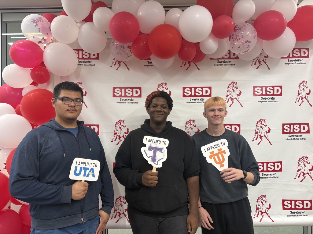 Three young people stand before a banner and balloons. Each holds a sign, one with "UTA" and the others with a letter "T".