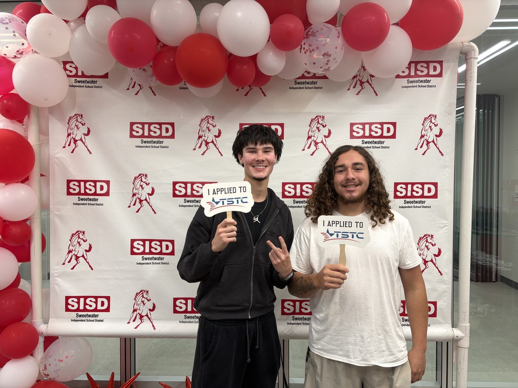 Two students pose for a photo with a photobooth backdrop of balloons and banners. They hold signs.