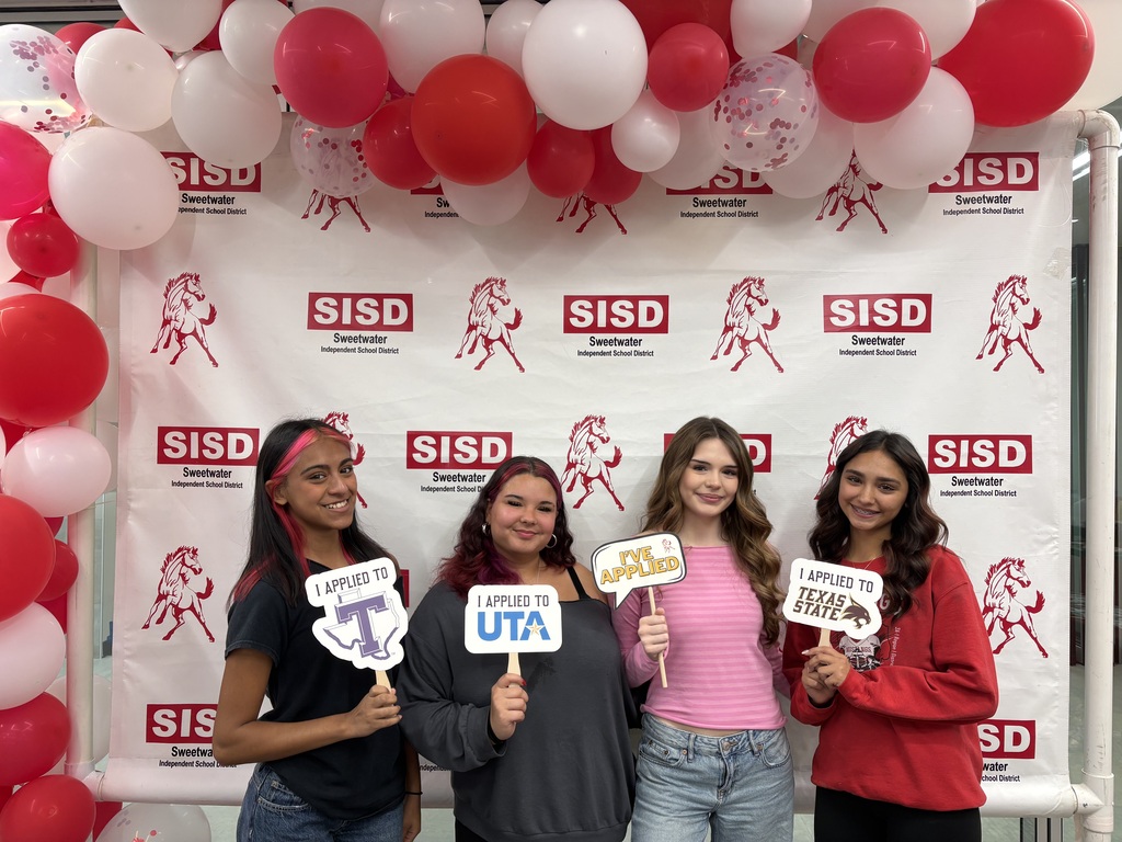 Four young women stand in front of a banner with red and white balloons, holding signs.