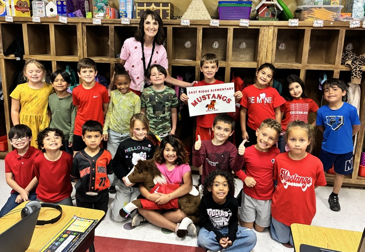 A classroom with children and a woman standing; one child holds a sign that says Mustangs.