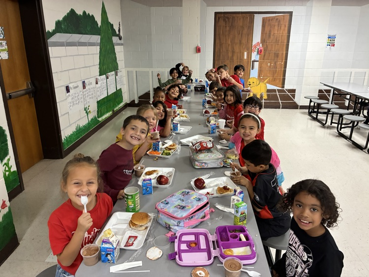 Children sit at long tables in a school cafeteria, eating food and drinks. Some hold utensils.