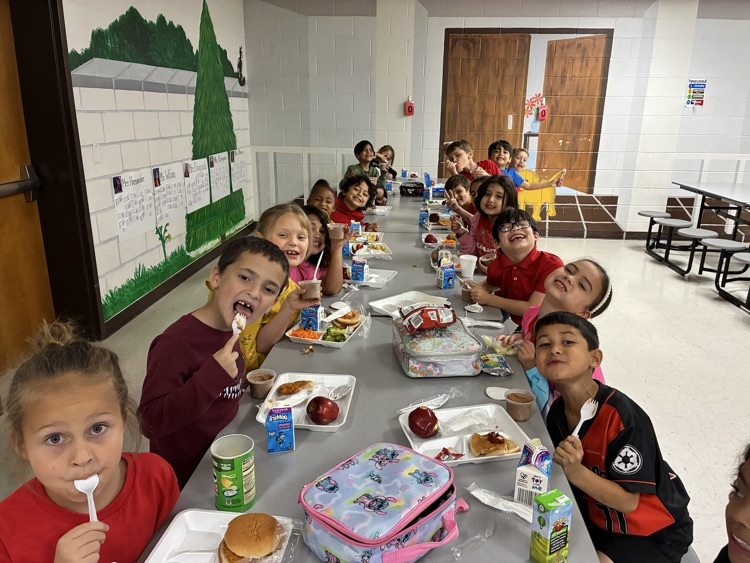 Students seated at tables in a cafeteria with trays of food, cups, and utensils.