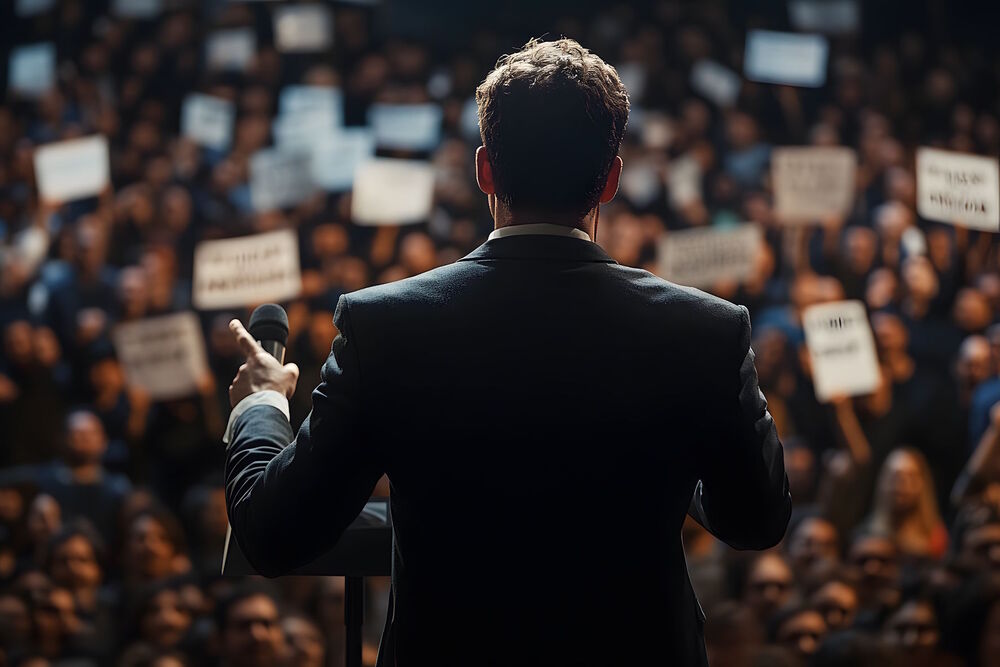 Man in a suit framed from behind giving a rousing political speech to a crowd of people who are holding signs.