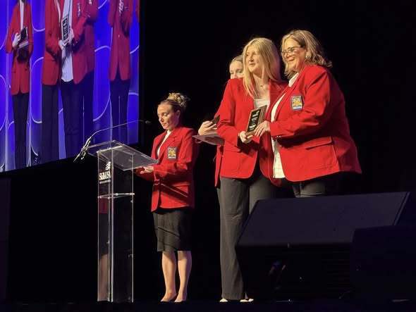 Four women stand on a stage, with two of them holding an award. The woman in the center wears a red jacket and holds an award, while the others are in red jackets.