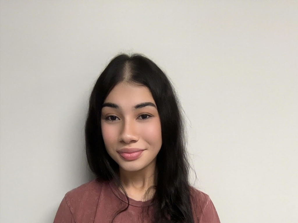 A woman with long black hair, smiling and wearing a maroon top, stands in front of a plain white background.