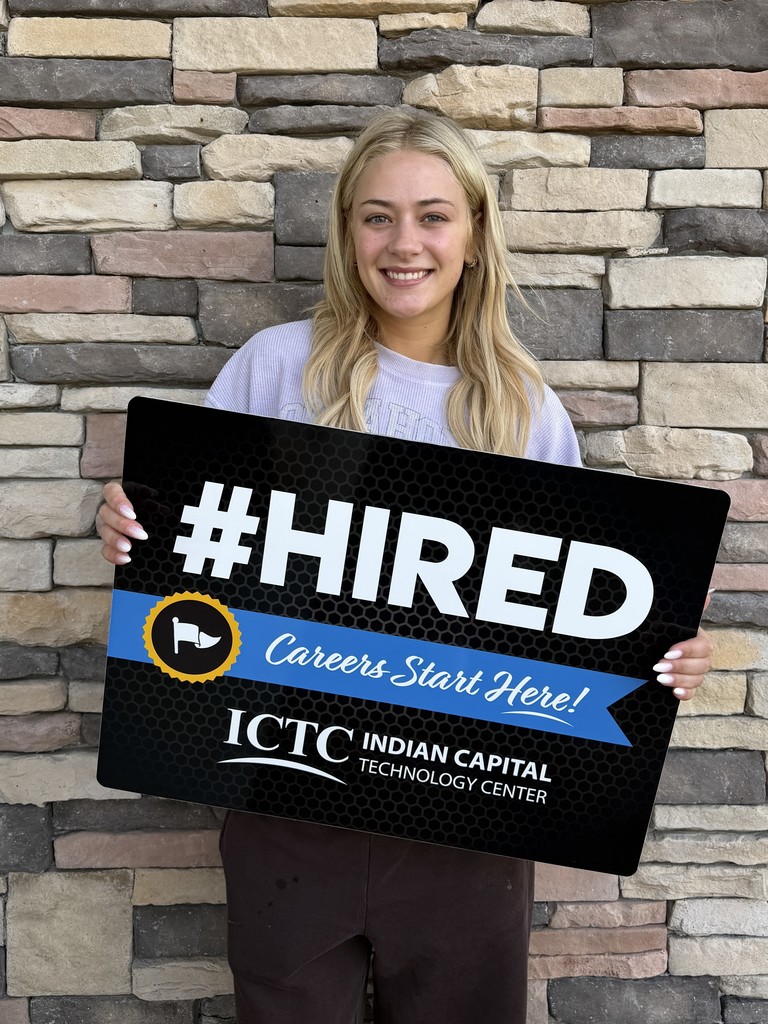 A woman holding a black sign reading "#HIRED" with a blue ribbon and logo for ICTC. She stands before a brick wall.
