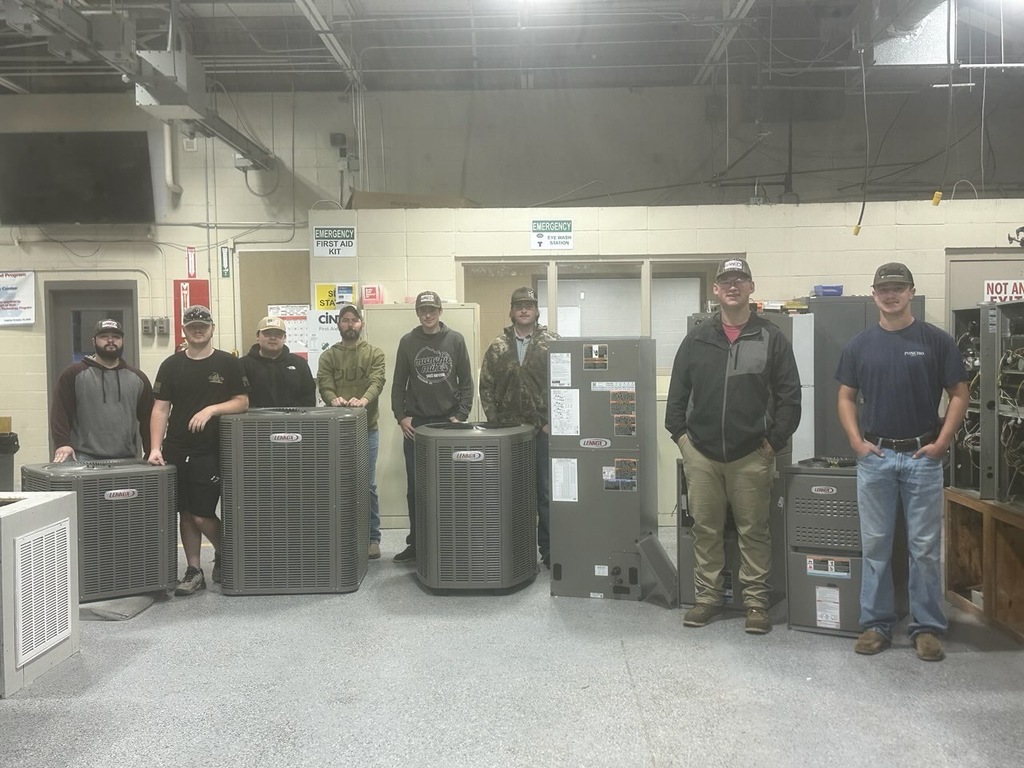 Nine men stand behind several HVAC units in a warehouse; some wear hats and sunglasses.