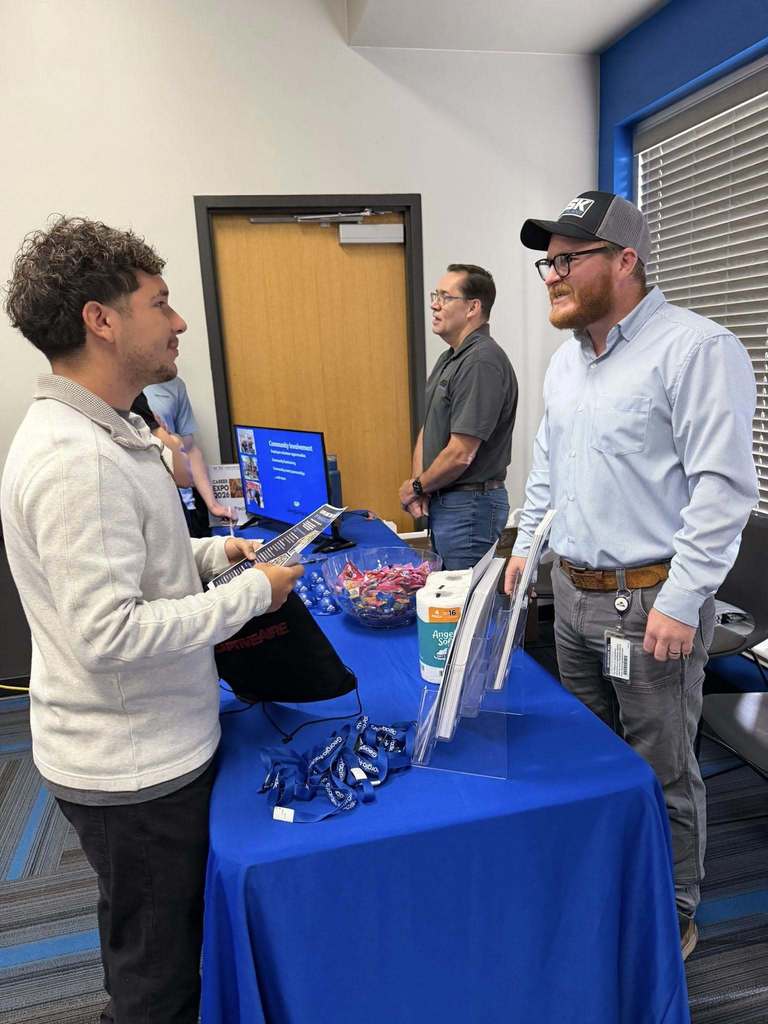 Two men at a table with a blue cover, one holding a tablet. Other men stand nearby, with a door and a monitor in the background.