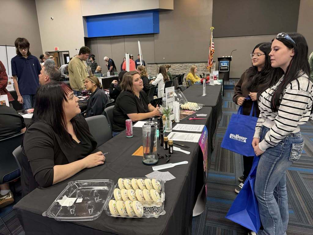 People in a room with tables. One person seated, one standing holding a bag. Food on a table.
