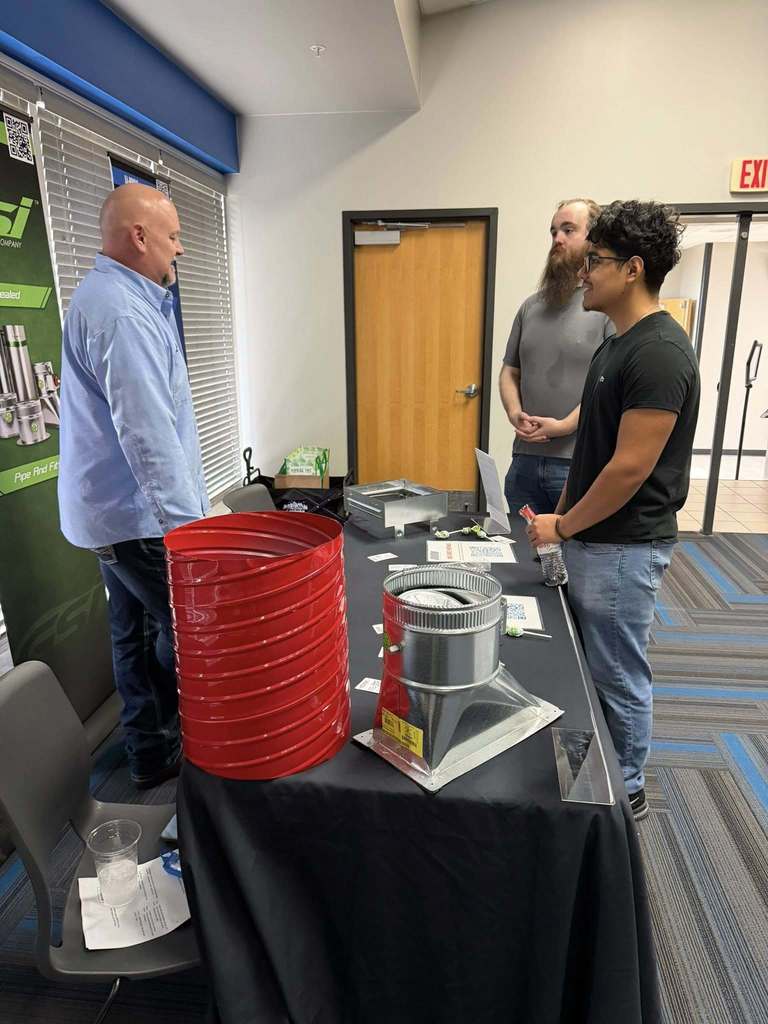 Three individuals at an event, one with a red cylinder, another with a silver cylinder, and a table with products.