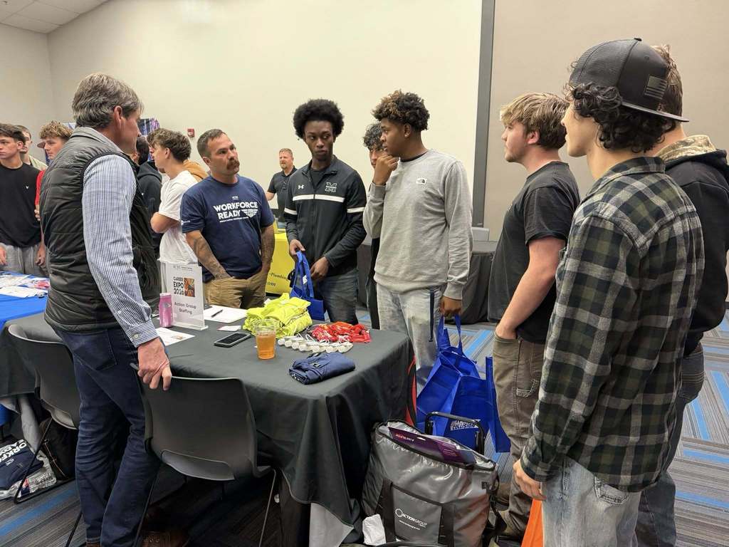 A group of people around a table with items, including backpacks. A man in a vest looks at the items.