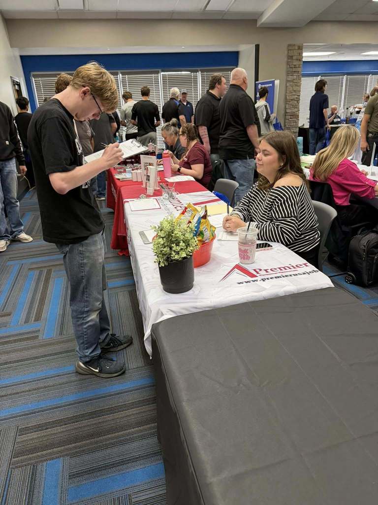 People at an event with tables. A man reads from a paper at a table. A woman sits at the same table.