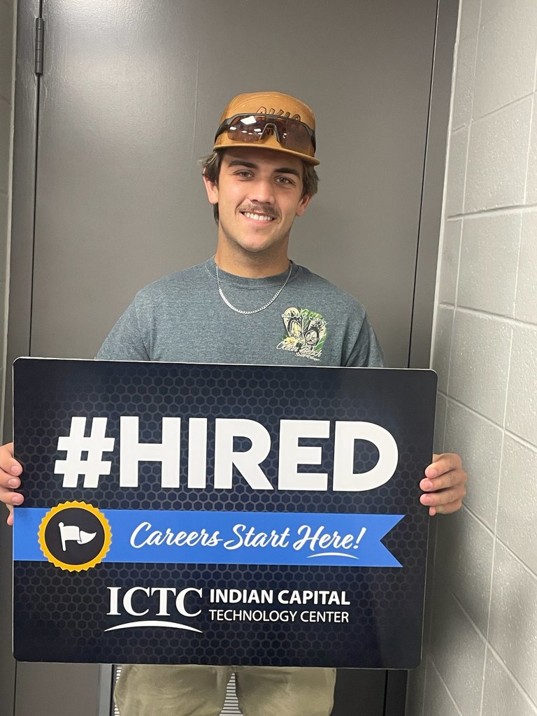 A man in sunglasses and a gray shirt holds a sign that reads "#HIRED" and "Careers Start Here!" in front of a tiled wall.